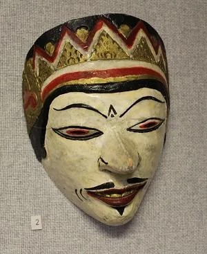 A traditional painted mask with a white face, black eyebrows, and red lips with gold, red, and black patterns.