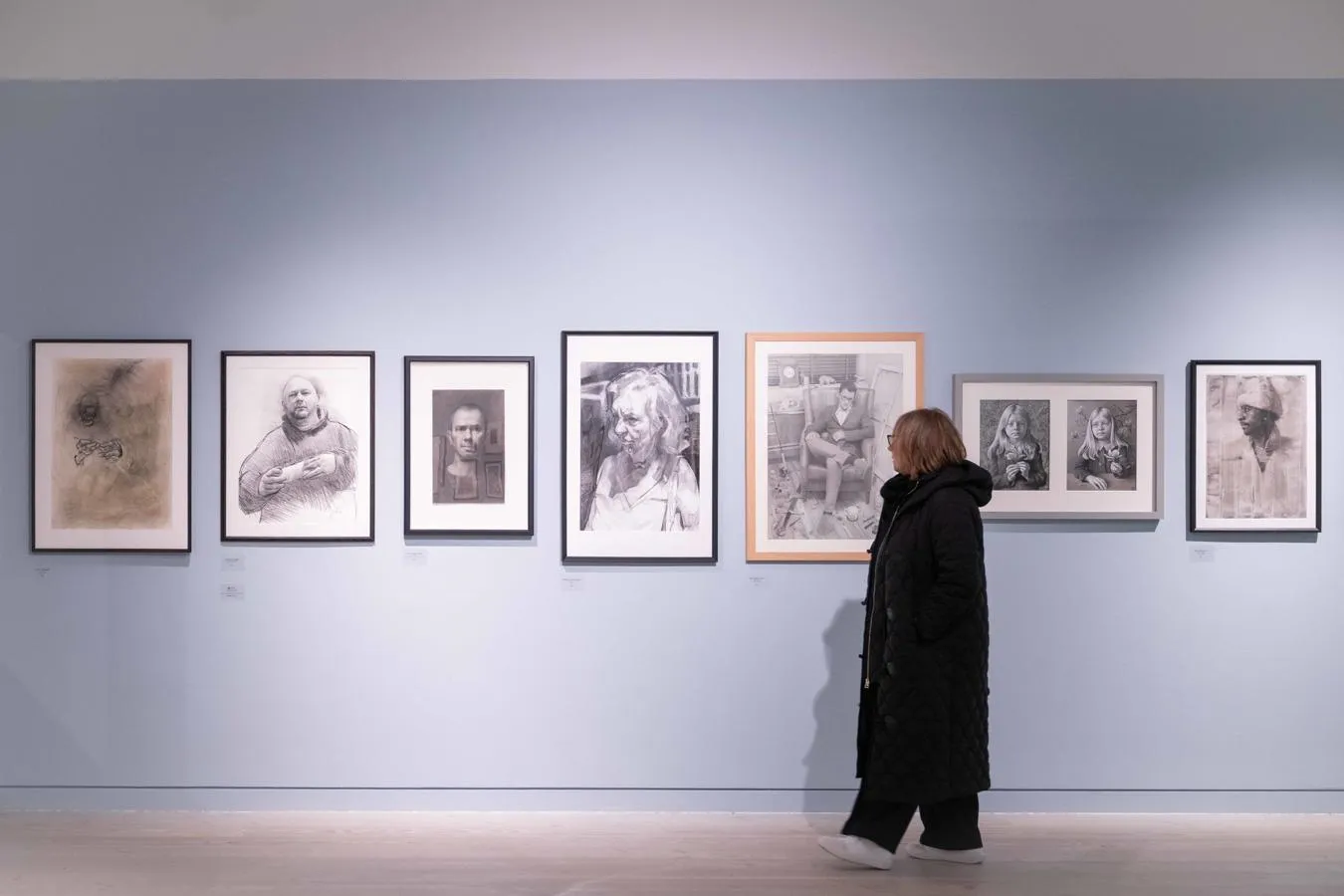 A person in a dark coat views a gallery wall displaying eight framed sketches and portraits.