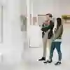 A man and a woman stand in a bright, minimalist art gallery, engaging with the exhibits.