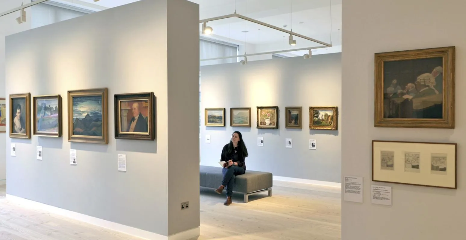 A person sits on a bench in an art gallery with light grey walls, admiring framed paintings.