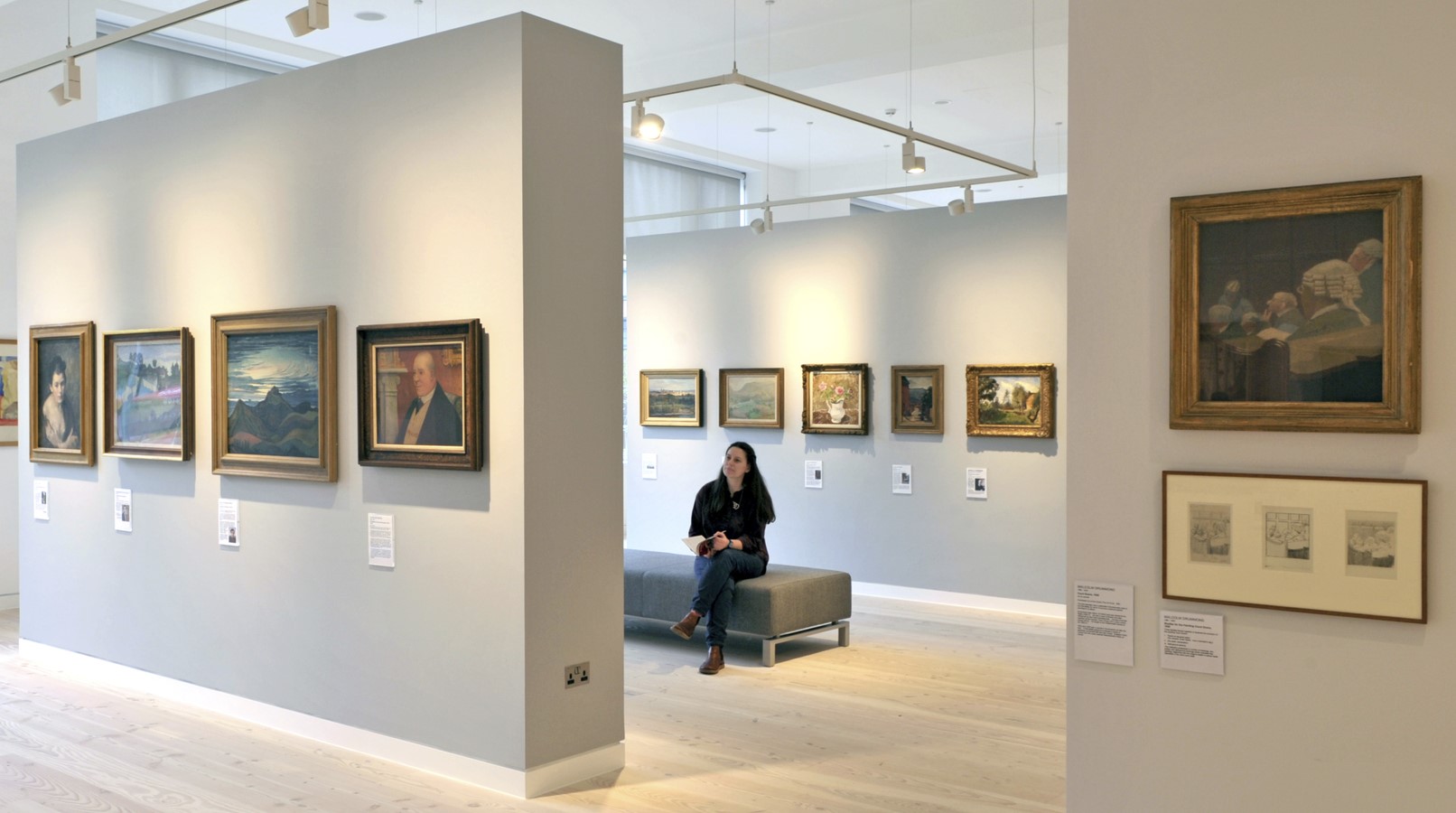 A person sits on a bench in an art gallery with light grey walls, admiring framed paintings. 